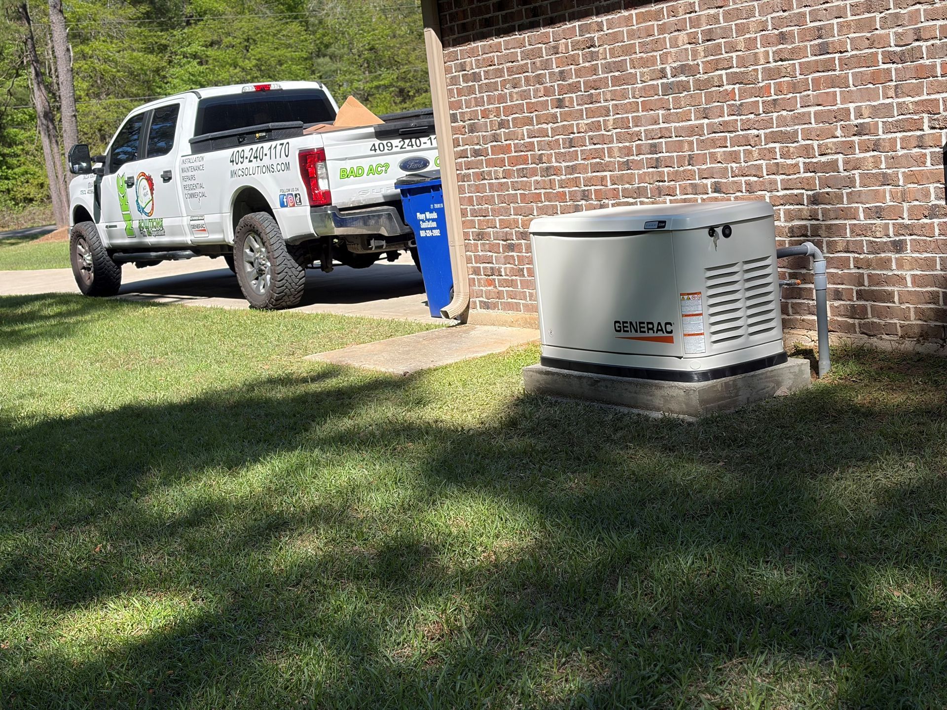 A white service truck is parked on a lawn next to a brick house with a Generac home standby generator on a concrete pad.
