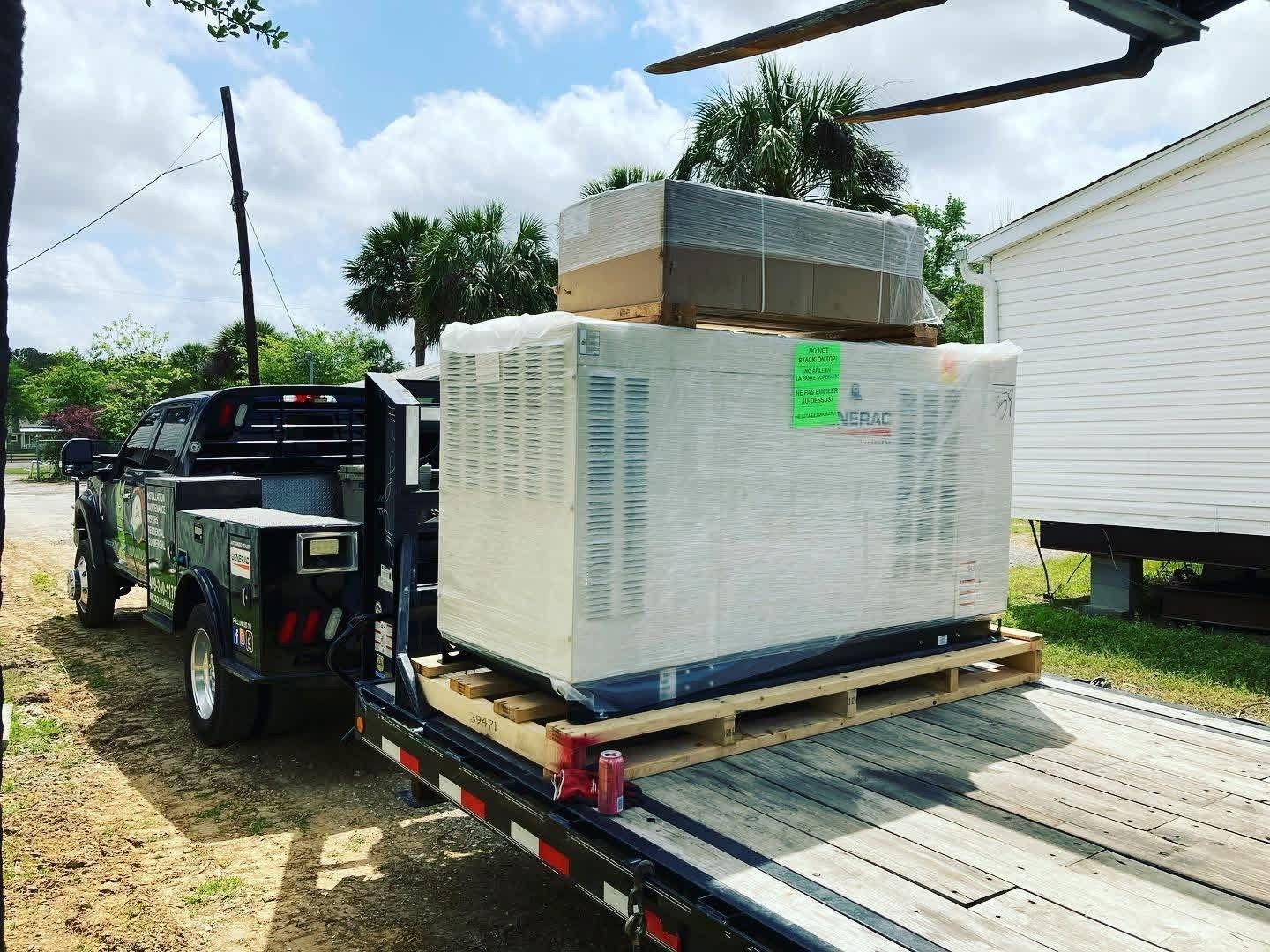 A black work truck pulling a flatbed trailer loaded with a large, wrapped pallet of materials next to a white house.