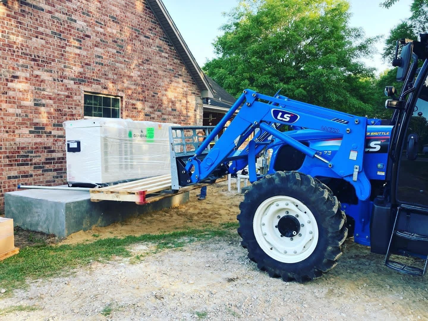 A blue tractor with a front loader is positioning a white generator onto a concrete slab next to a brick house.
