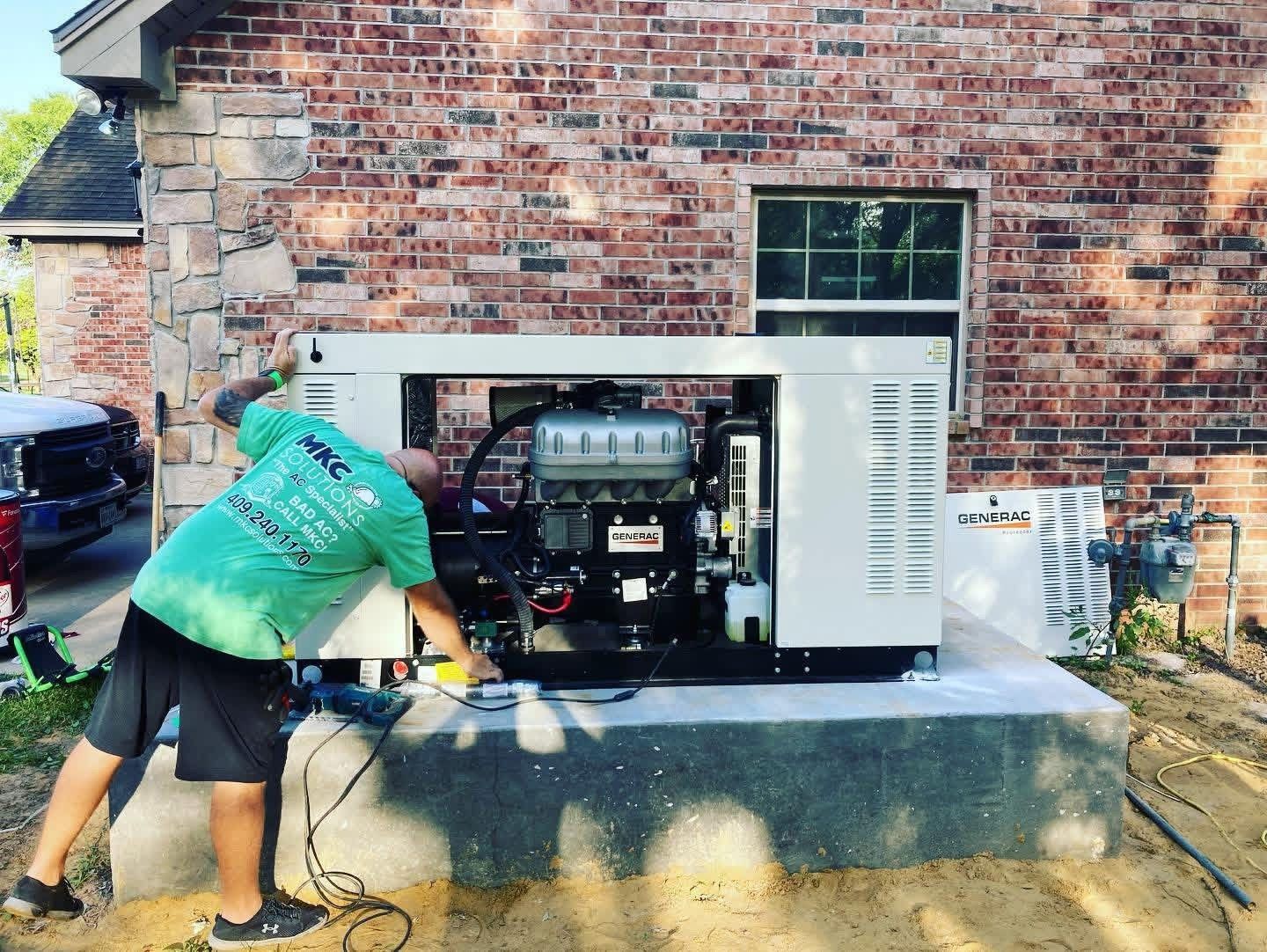 A person in a green t-shirt working on a residential backup generator installed on a concrete pad outside a brick house.