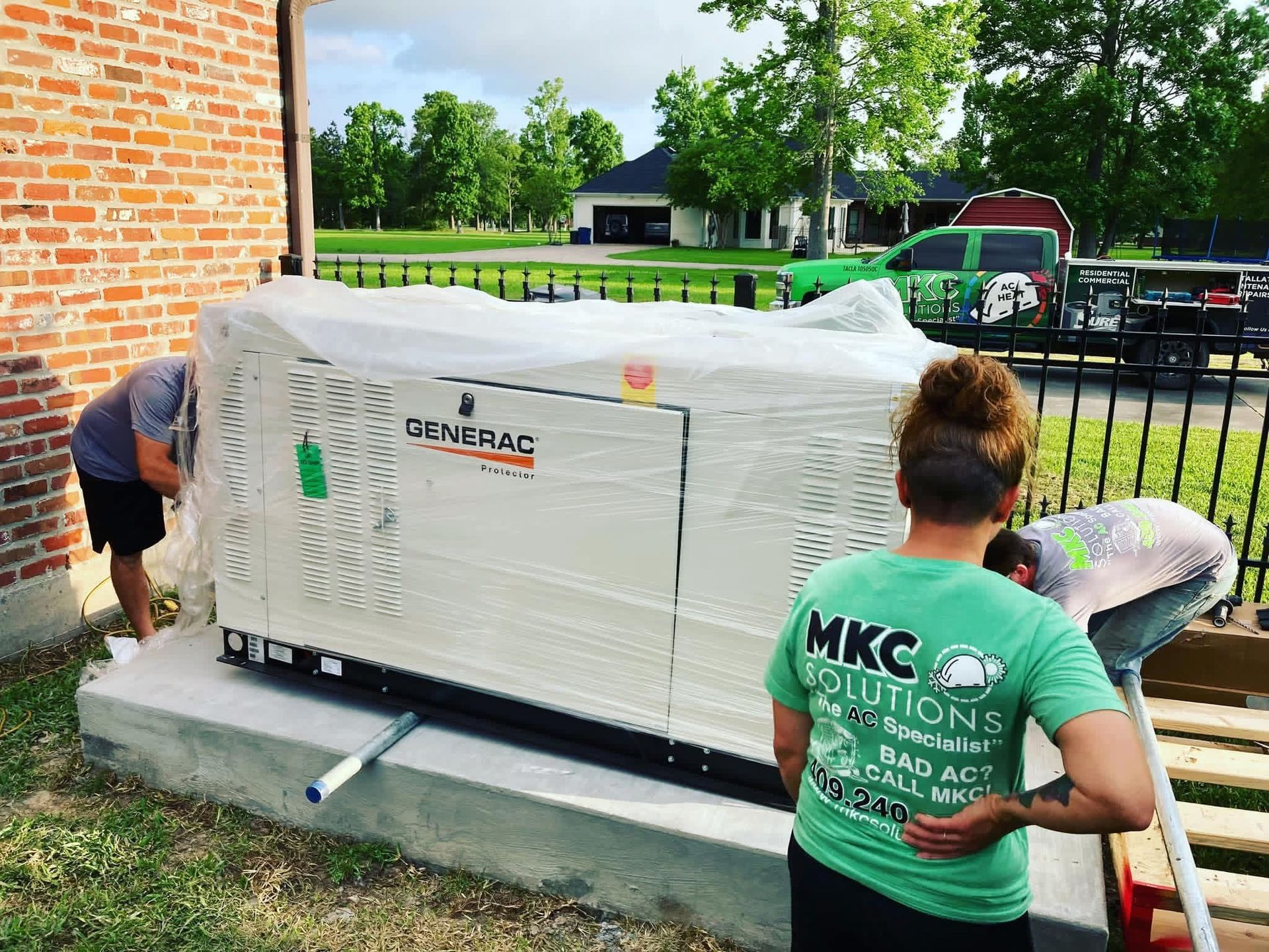 Workers install a Generac backup generator on a concrete pad next to a brick house.
