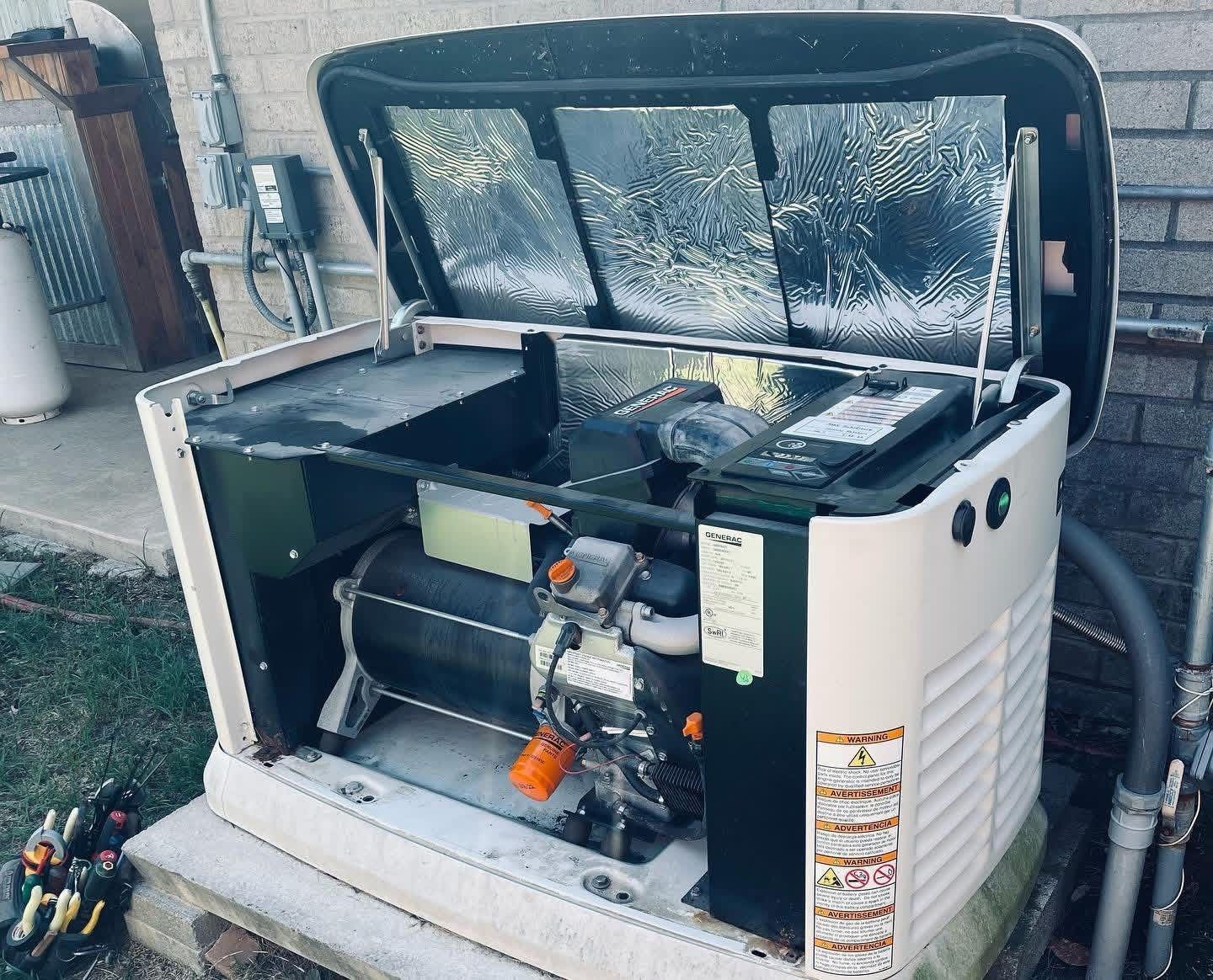 A standby home generator with its lid propped open, situated on a concrete pad outside a brick building.