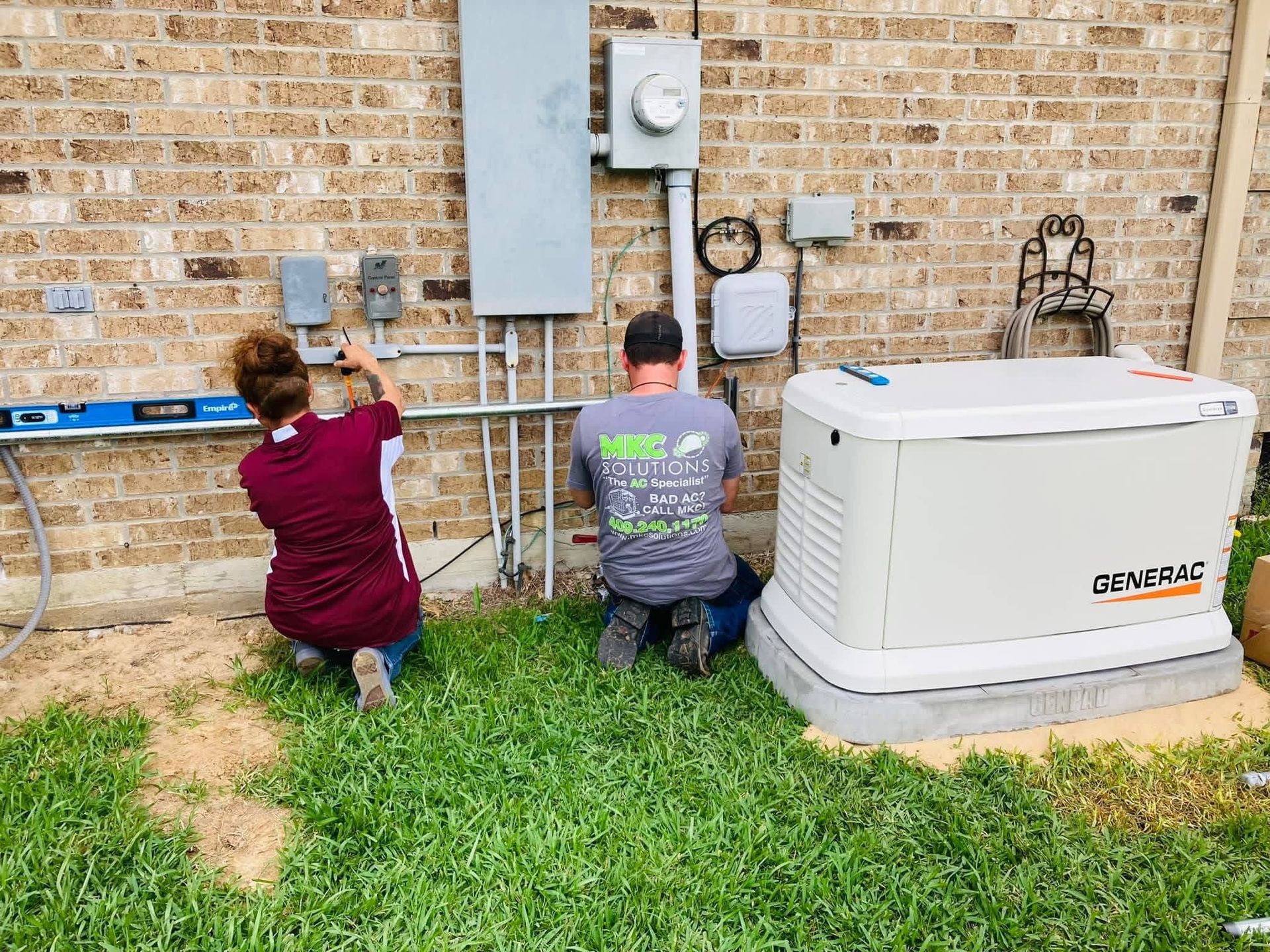 Two workers kneel by a brick wall, installing electrical equipment next to a large, white standby home generator.