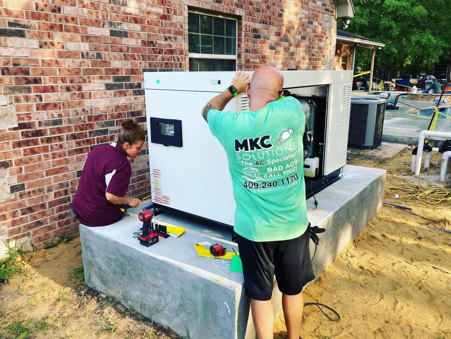 Two people work on a white outdoor generator installed on a concrete pad next to a brick house.