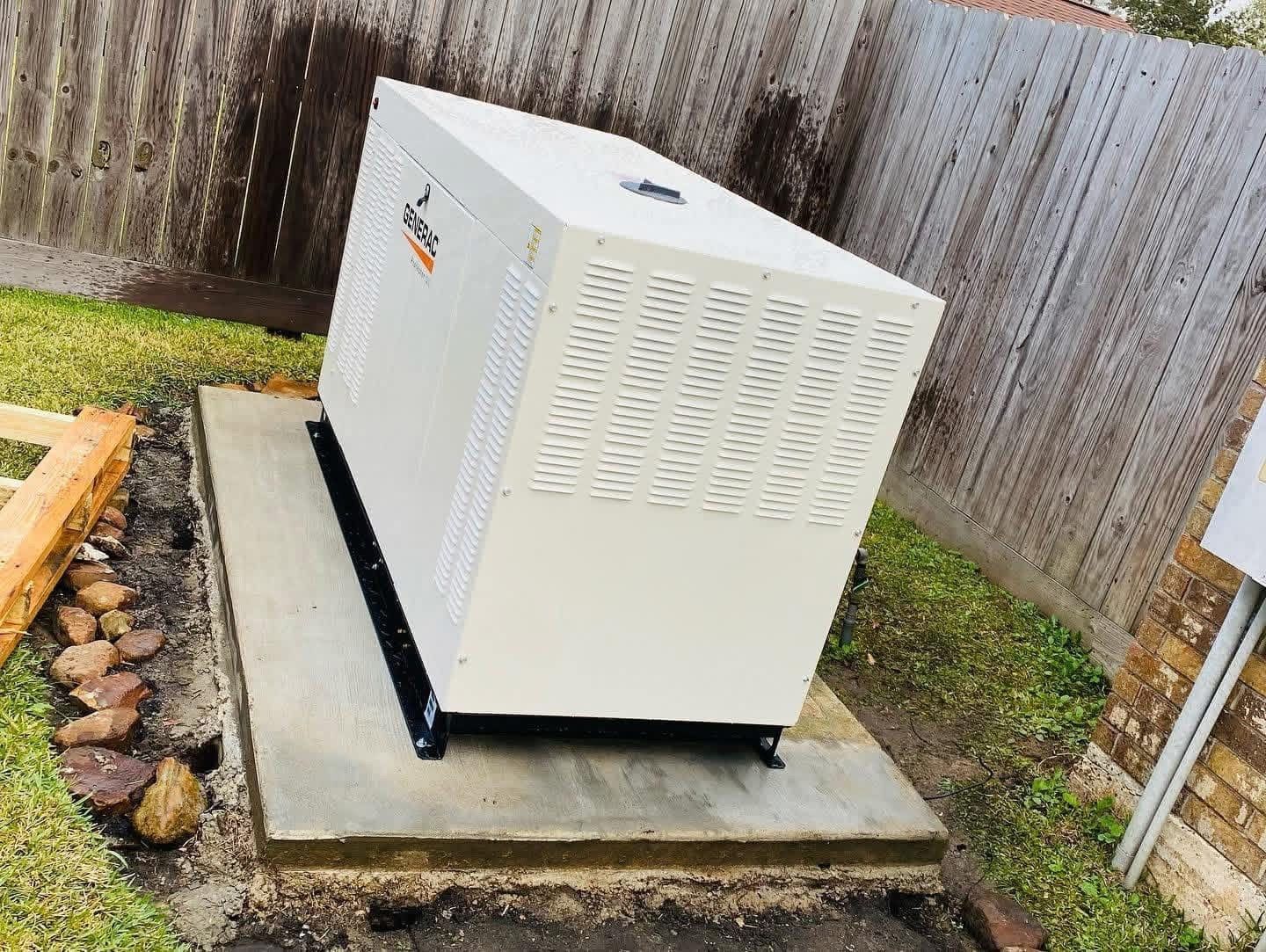 A white backup home generator sits on a concrete pad in a backyard next to a wooden fence.