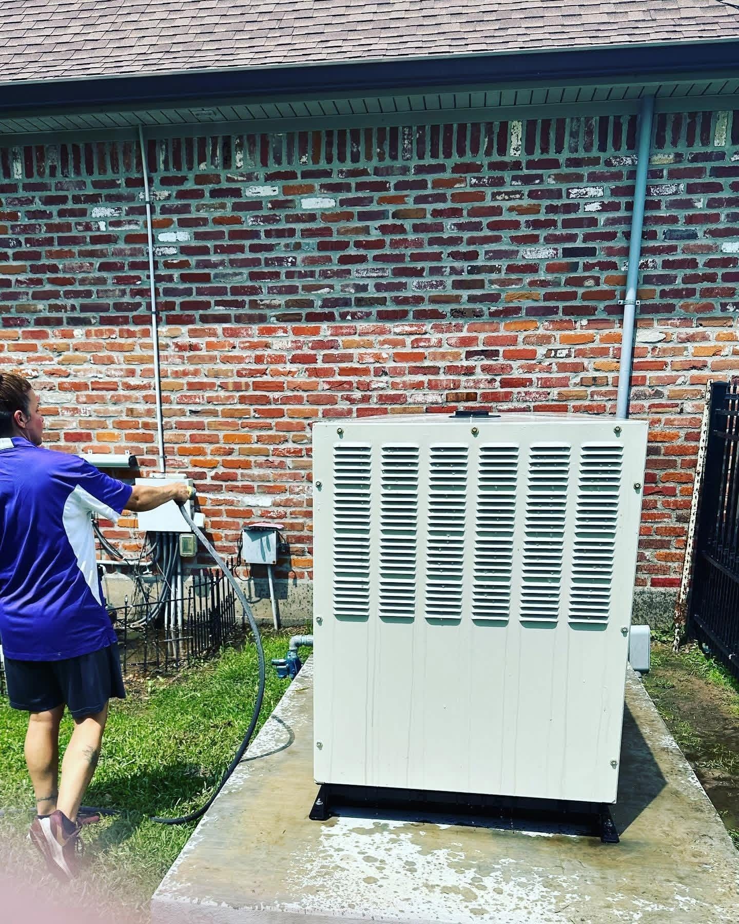 A person in a purple shirt uses a power washer to clean a beige generator sitting on a concrete pad against a brick wall.