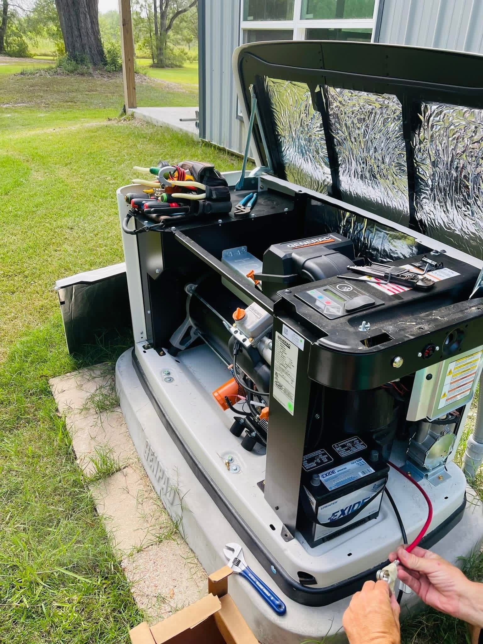 An open residential standby generator outdoors with tools nearby and hands connecting a red battery cable.