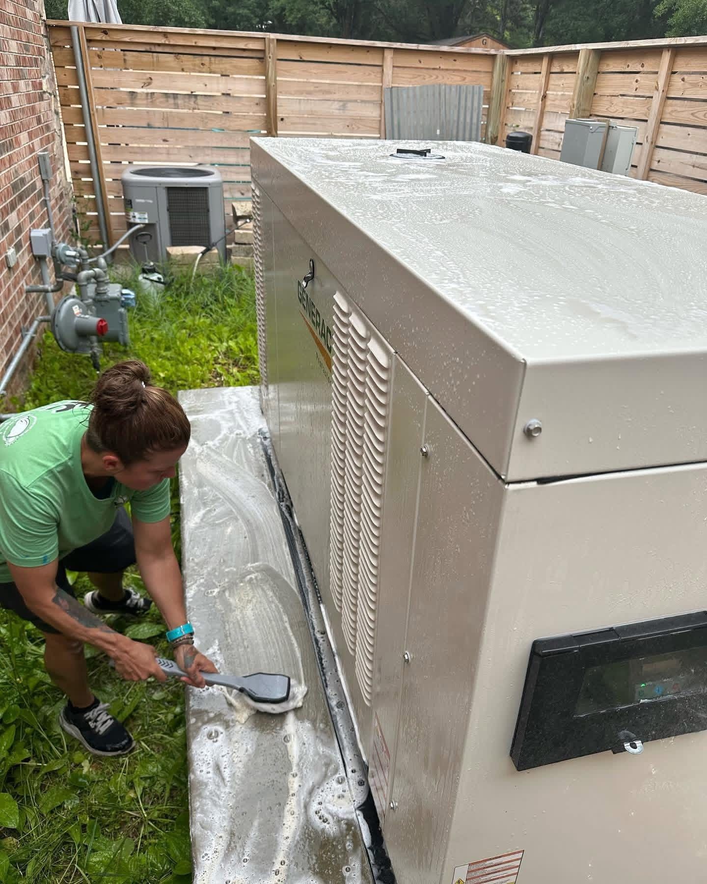 A person in a green shirt scrubs the concrete base of an outdoor backup generator with a long-handled brush.