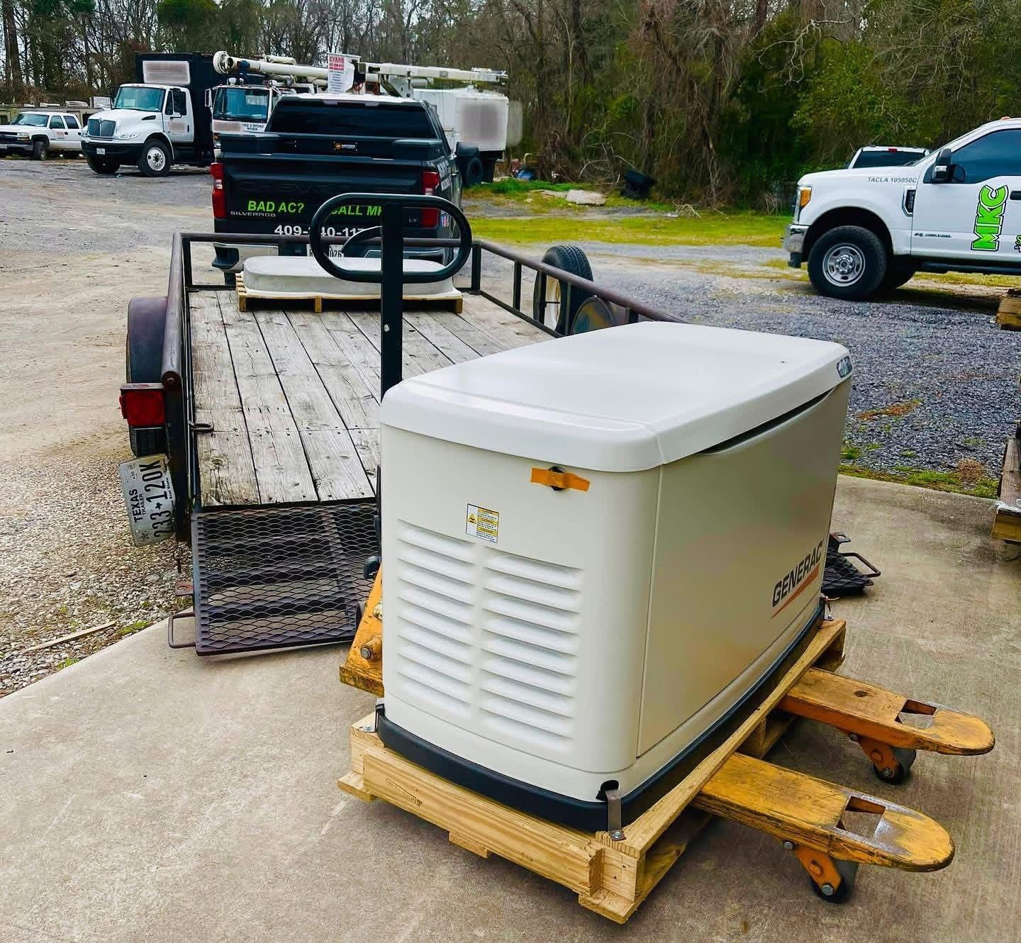 A beige generator on a wooden pallet jack sits on a trailer attached to a black pickup truck in an outdoor lot.