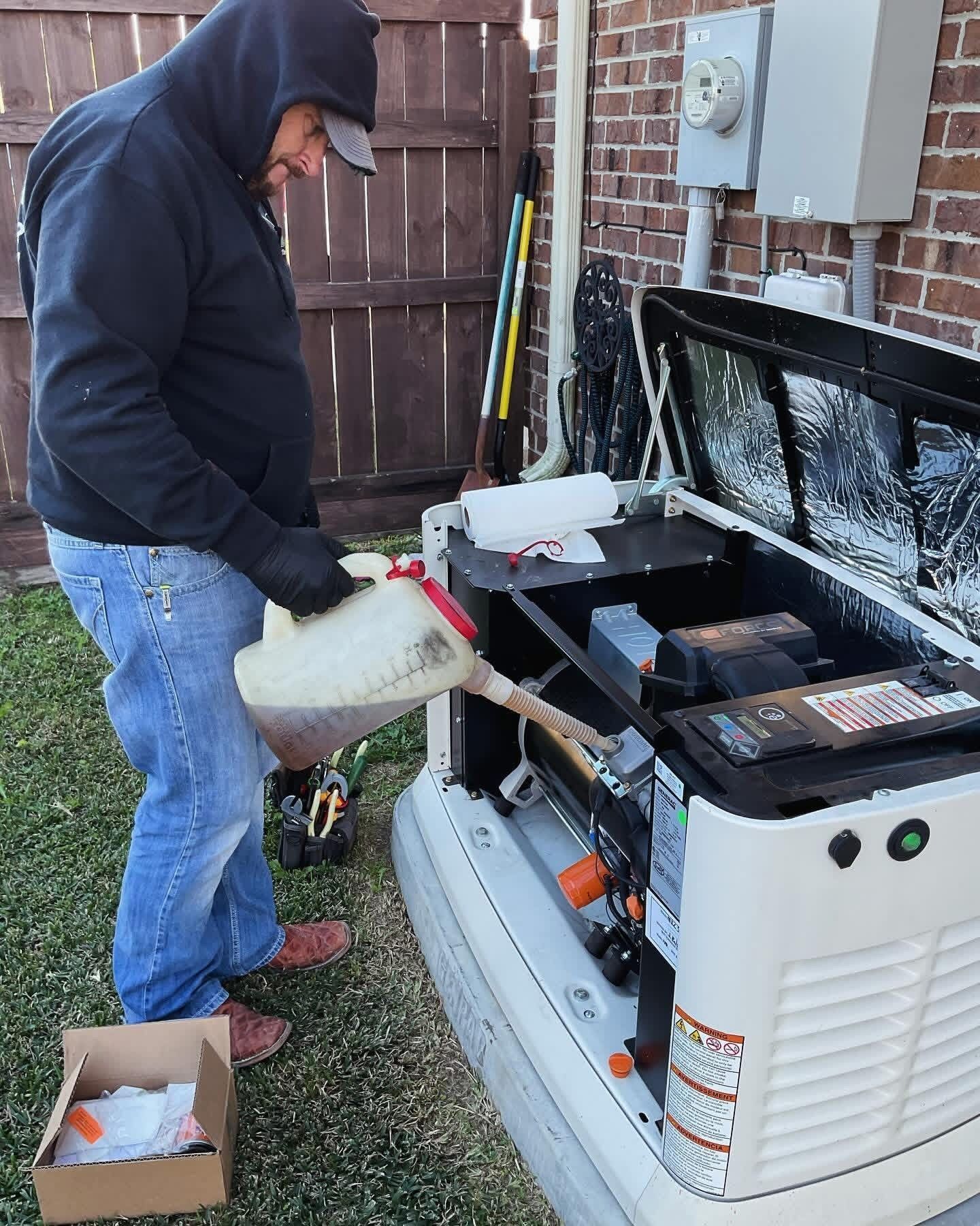 A person wearing a hoodie and gloves pours oil into a white residential backup generator outdoors next to a brick wall.