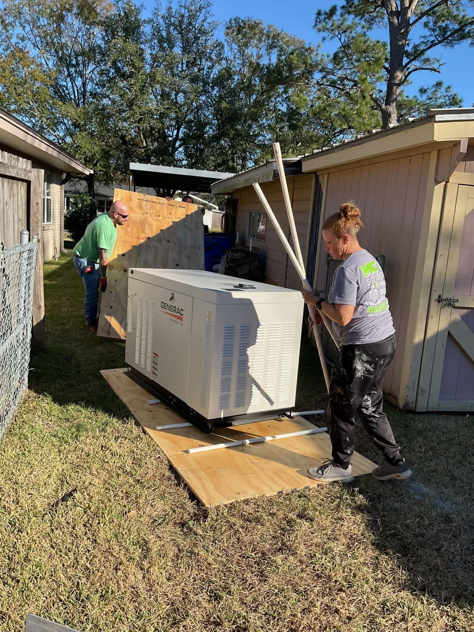 Two people move a white generator across a plywood sheet on a grassy residential lawn near a shed.