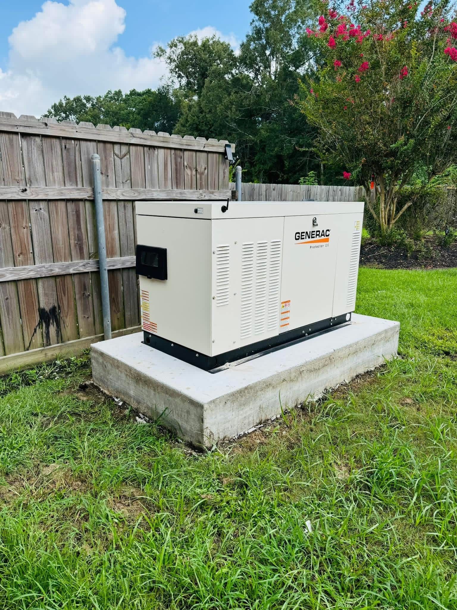 A cream-colored residential standby generator sits on a concrete pad in a grassy yard next to a wooden fence.