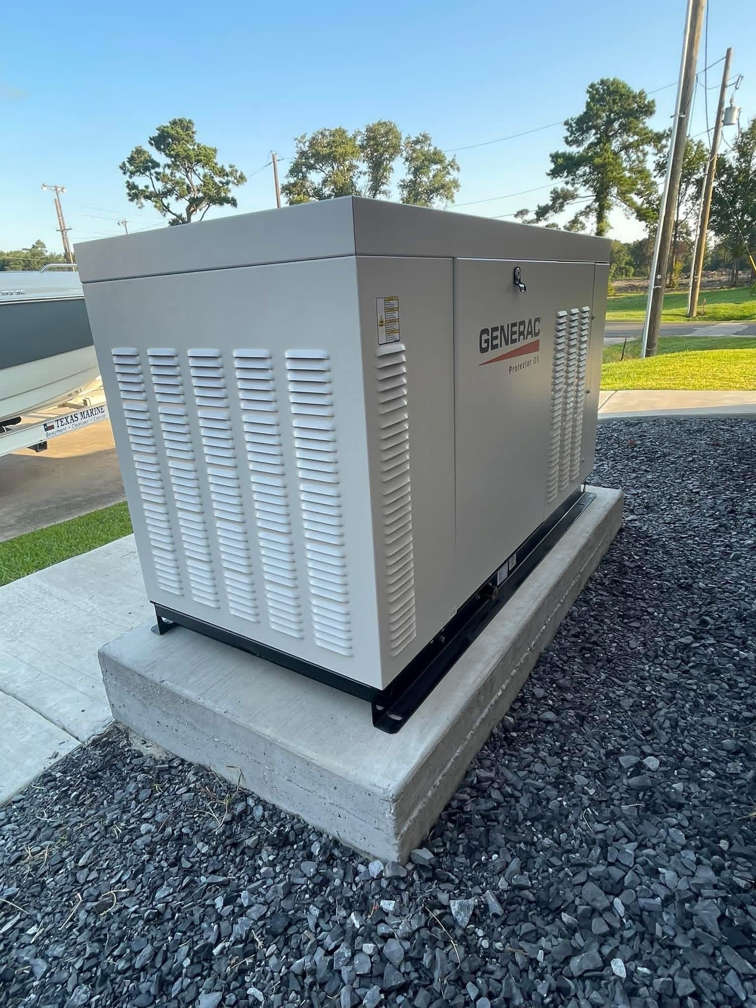 A Generac standby generator on a concrete pad surrounded by gravel in a grassy, outdoor setting.