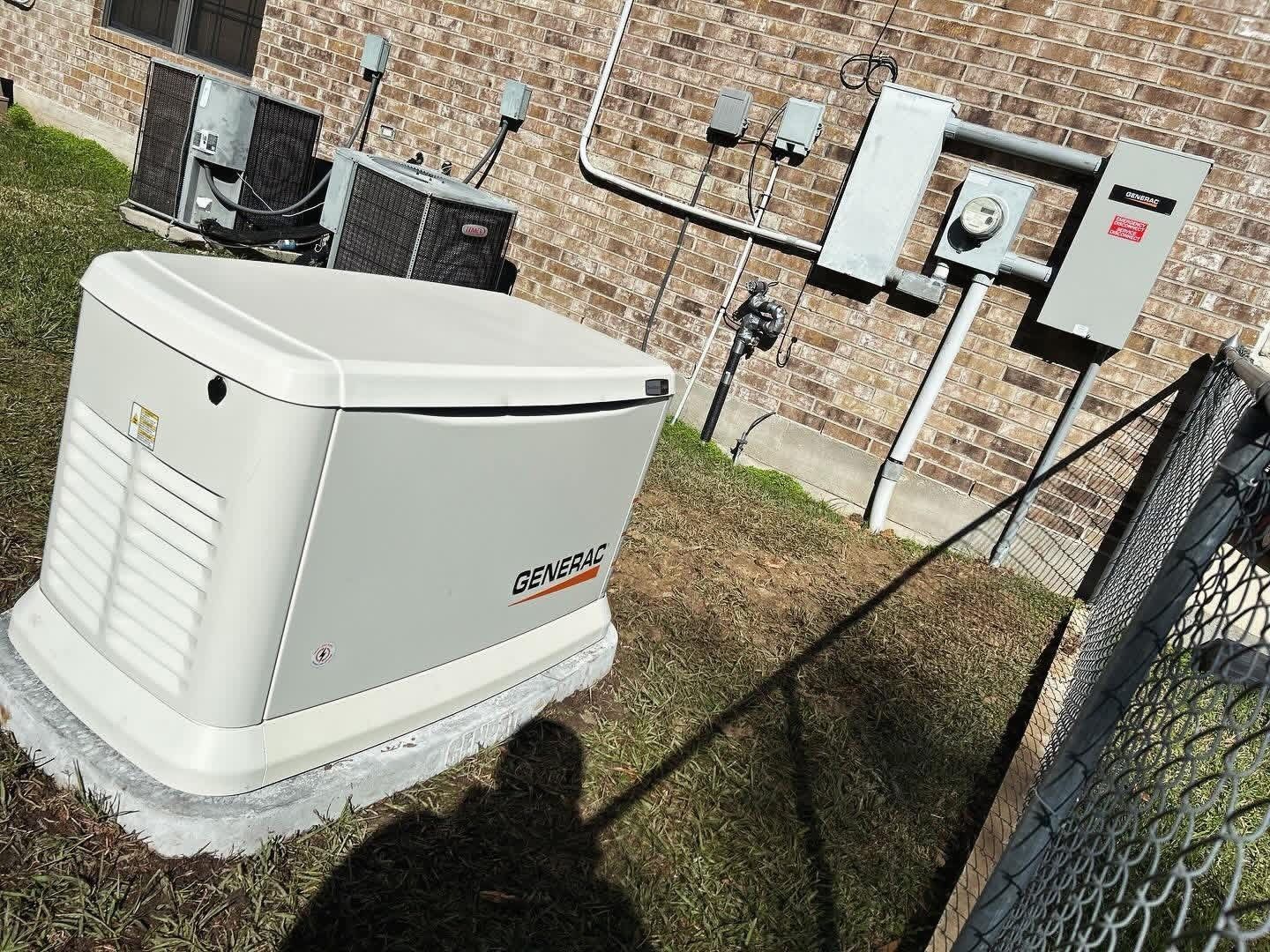 A Generac standby home generator sits on a concrete pad outside a brick house near electrical equipment and a chain fence.