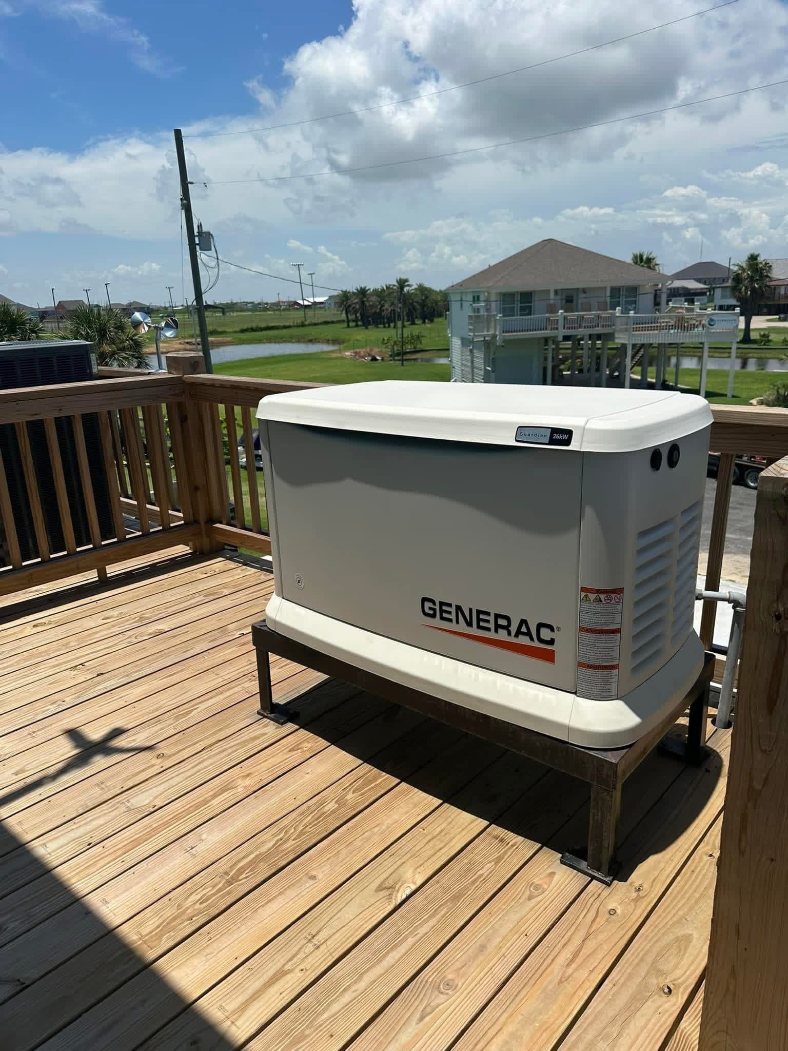 A Generac backup generator sits on a raised metal stand on a wooden deck overlooking a coastal area with houses.