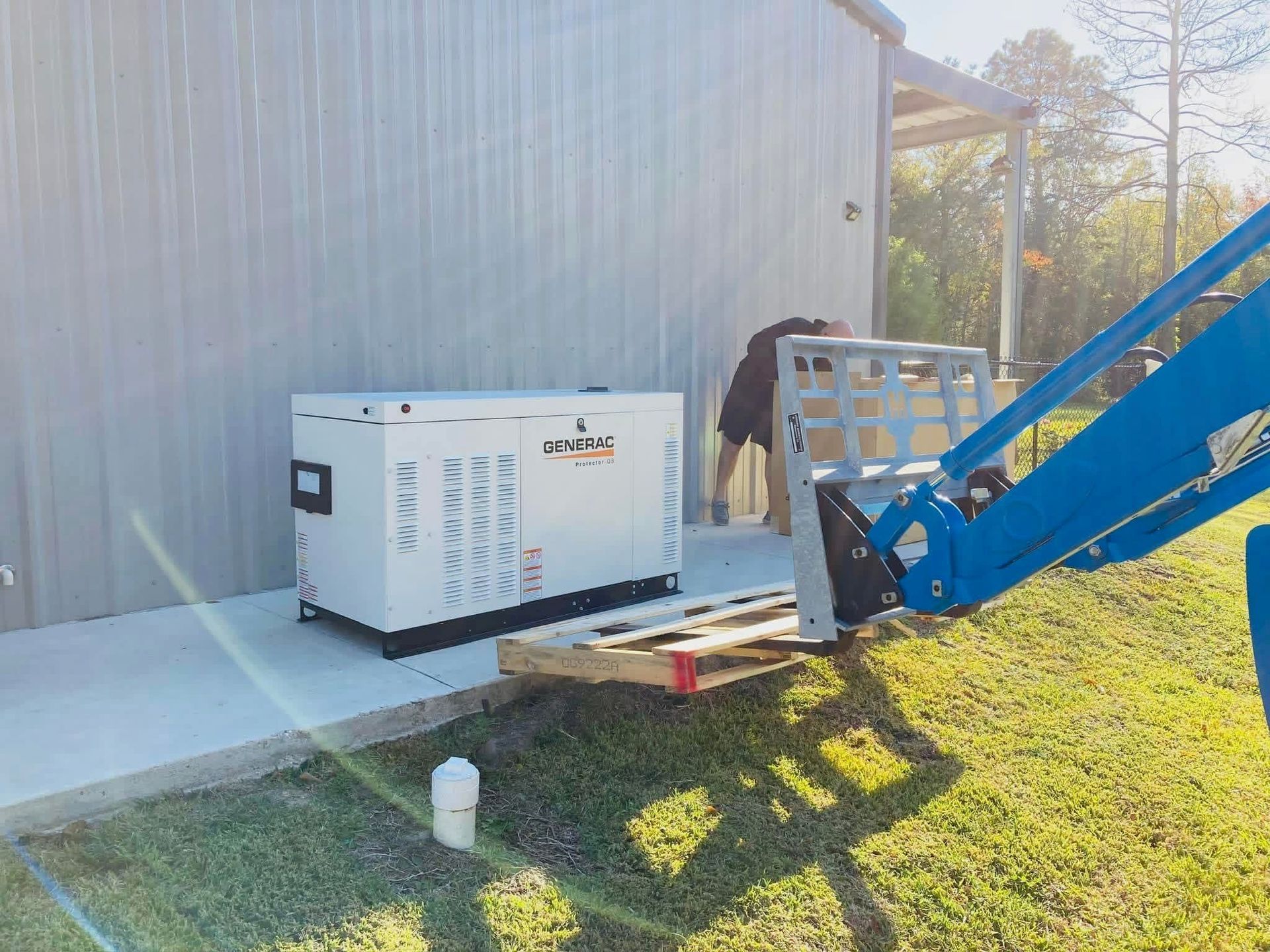 A white outdoor generator sits on a concrete pad next to a metal building as a blue forklift places a wooden pallet nearby.