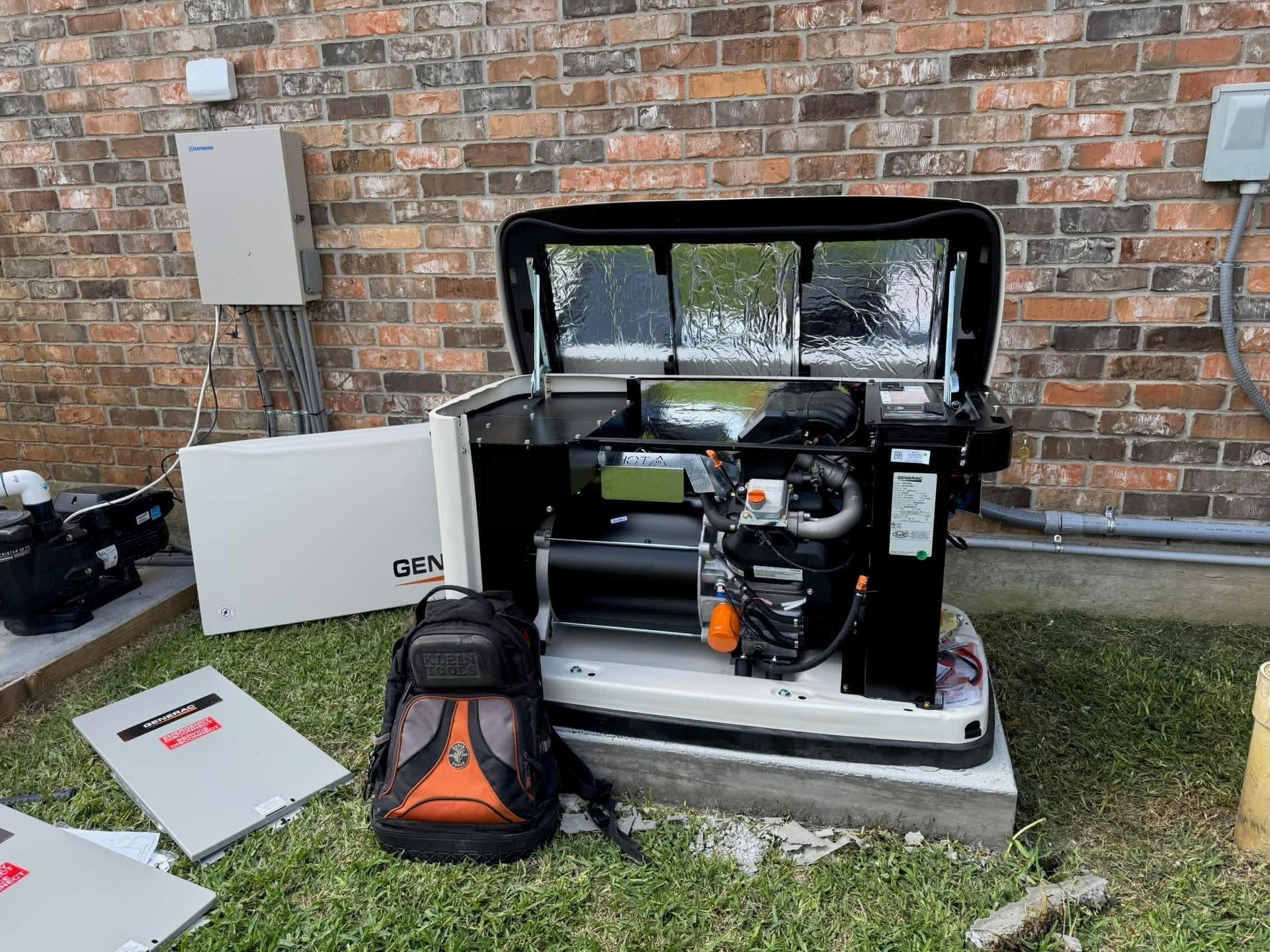 An open home standby generator sits on a concrete pad against a brick wall, with a backpack resting in front of it.