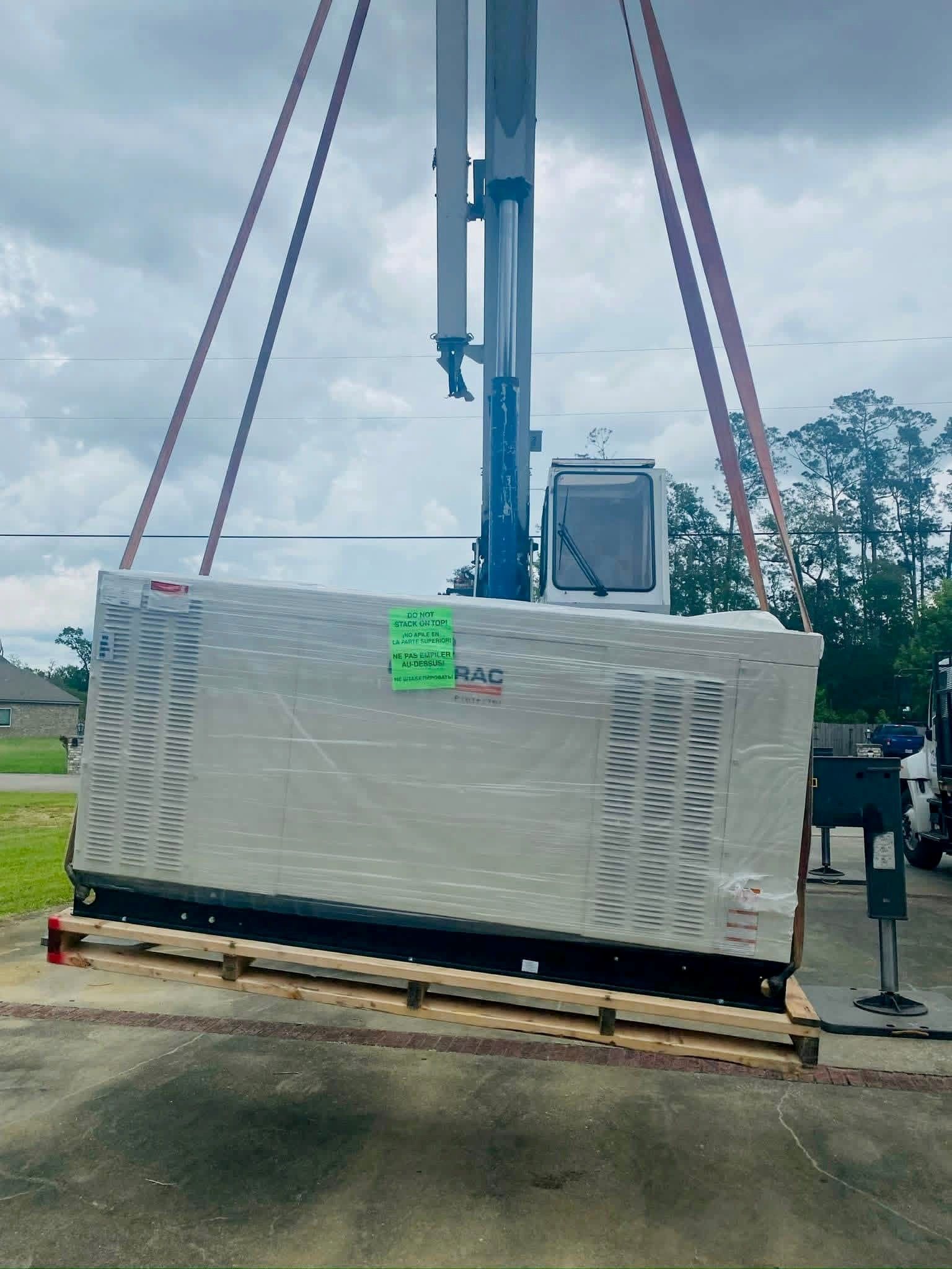 A large industrial generator wrapped in protective plastic is suspended in the air by a crane on a cloudy day.