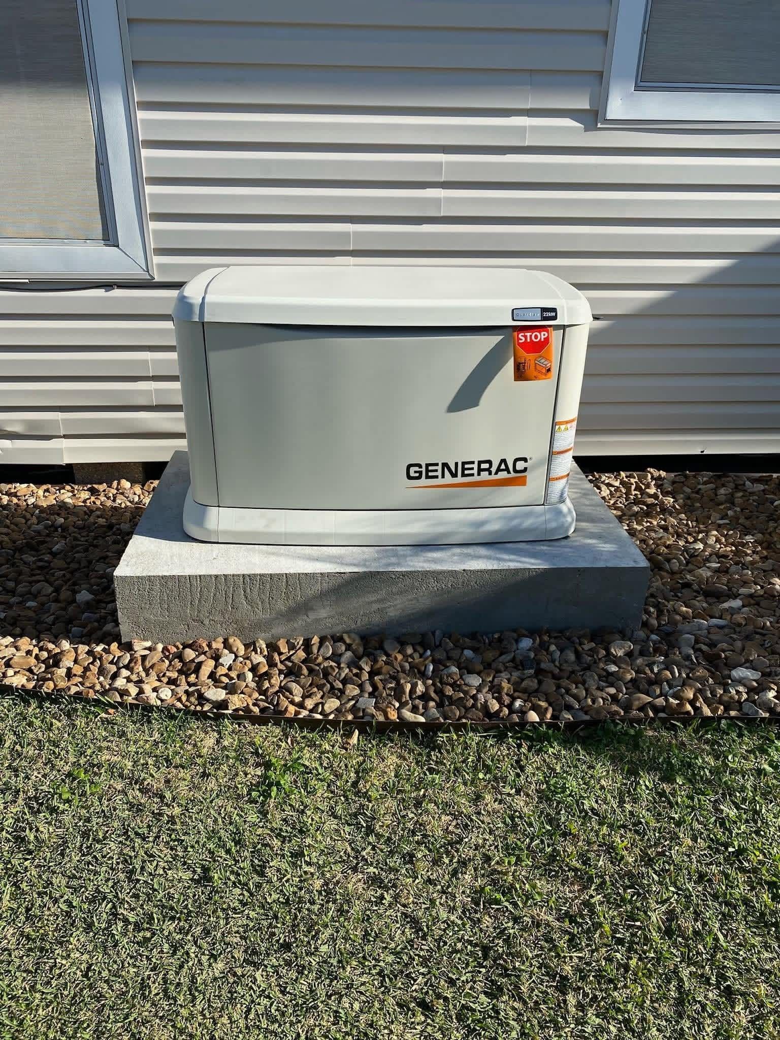 A grey Generac standby generator sitting on a concrete pad against white vinyl siding, surrounded by gravel and grass.