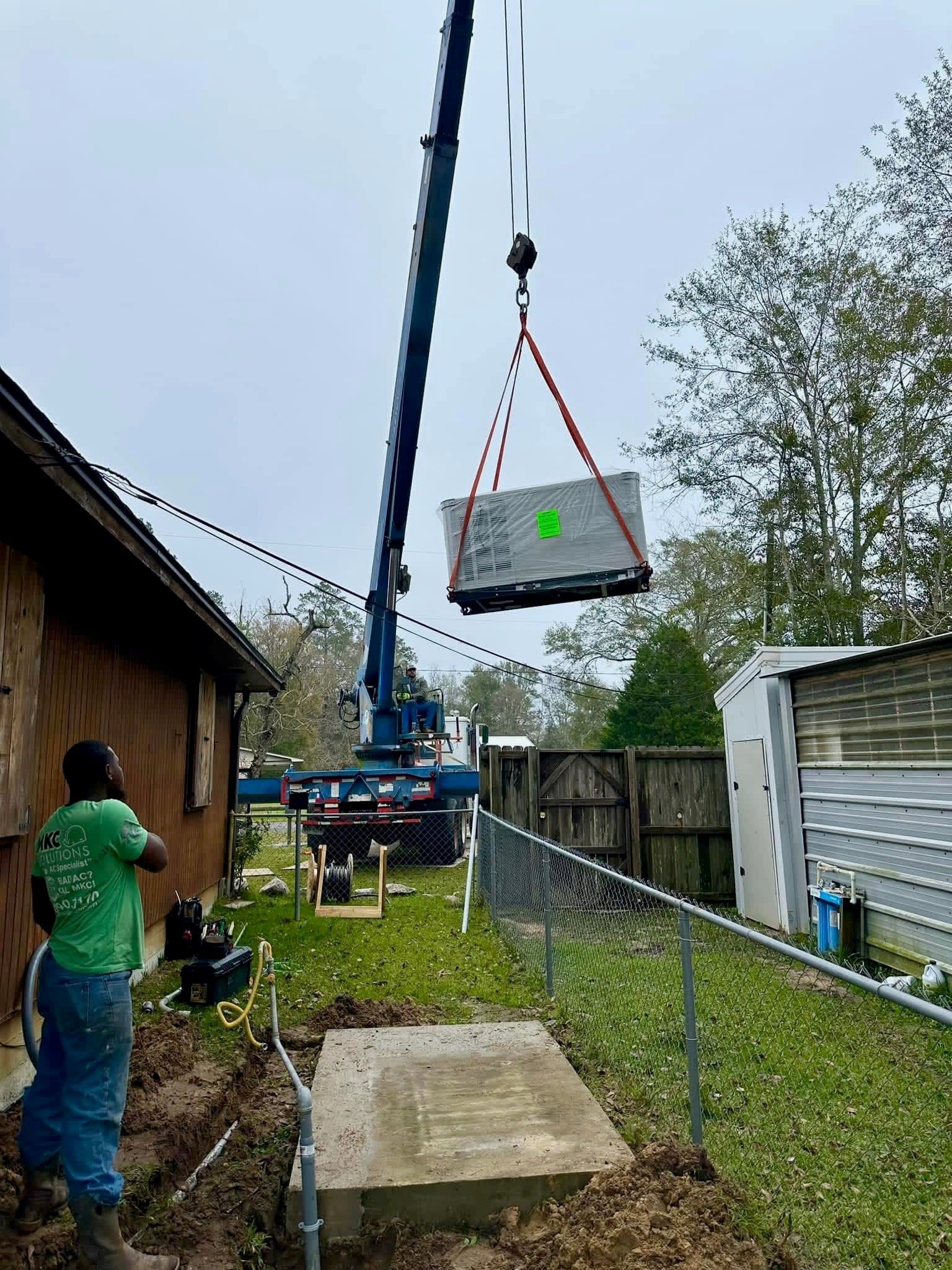 A crane lifts a large concrete slab over a yard toward a prepared foundation as a person watches the maneuver.