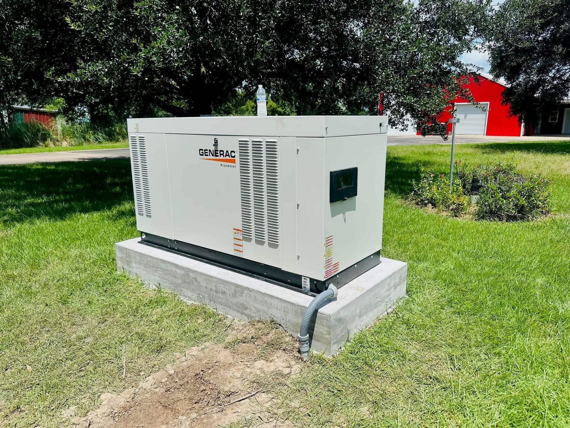 A beige residential standby generator sits on a concrete pad in a grassy yard, with trees and a red building in the back.