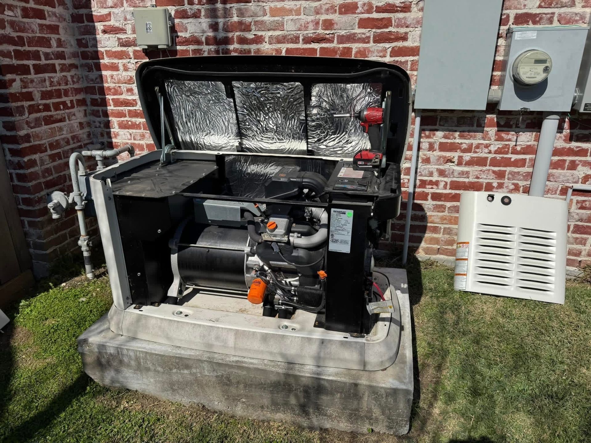 A standby home generator with its lid open, revealing the engine, sits on a concrete pad by a red brick exterior wall.