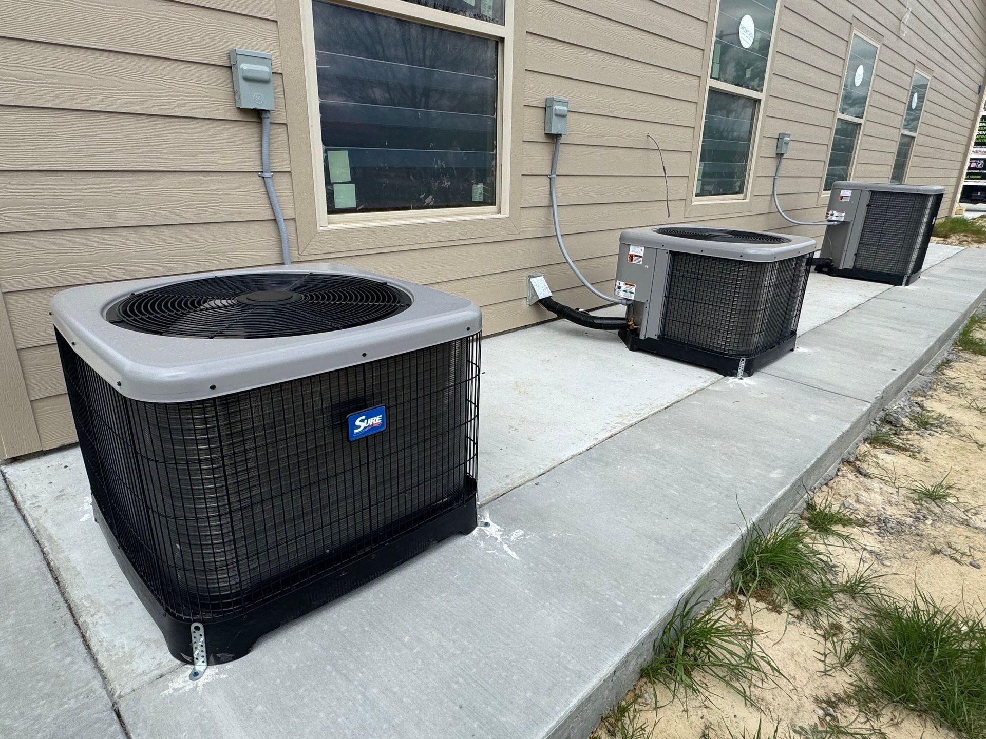 Three air conditioners are sitting on the sidewalk outside of a building.