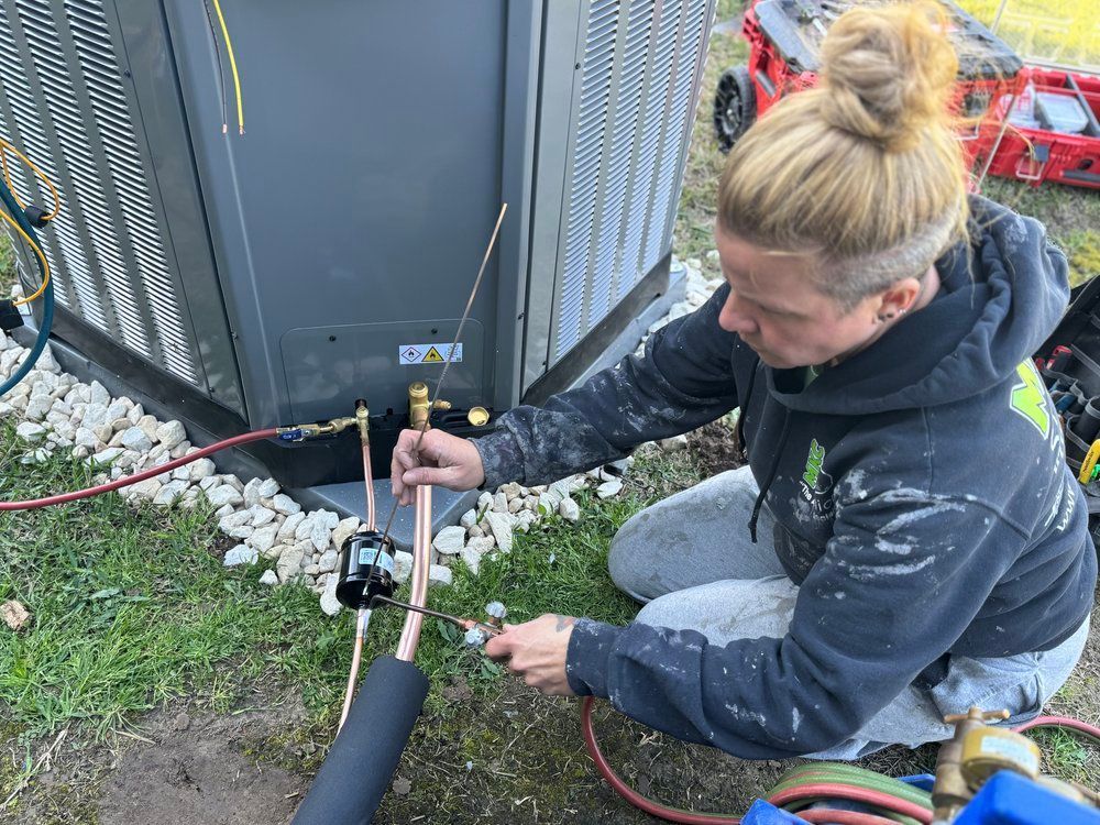 A woman is kneeling down in the grass working on an air conditioner.