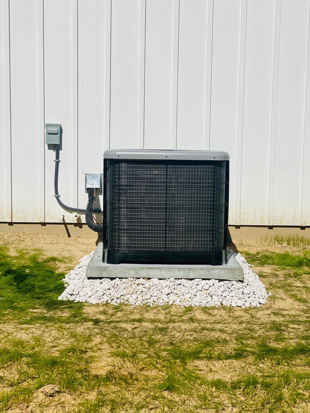 An air conditioner is sitting in the grass in front of a white fence.