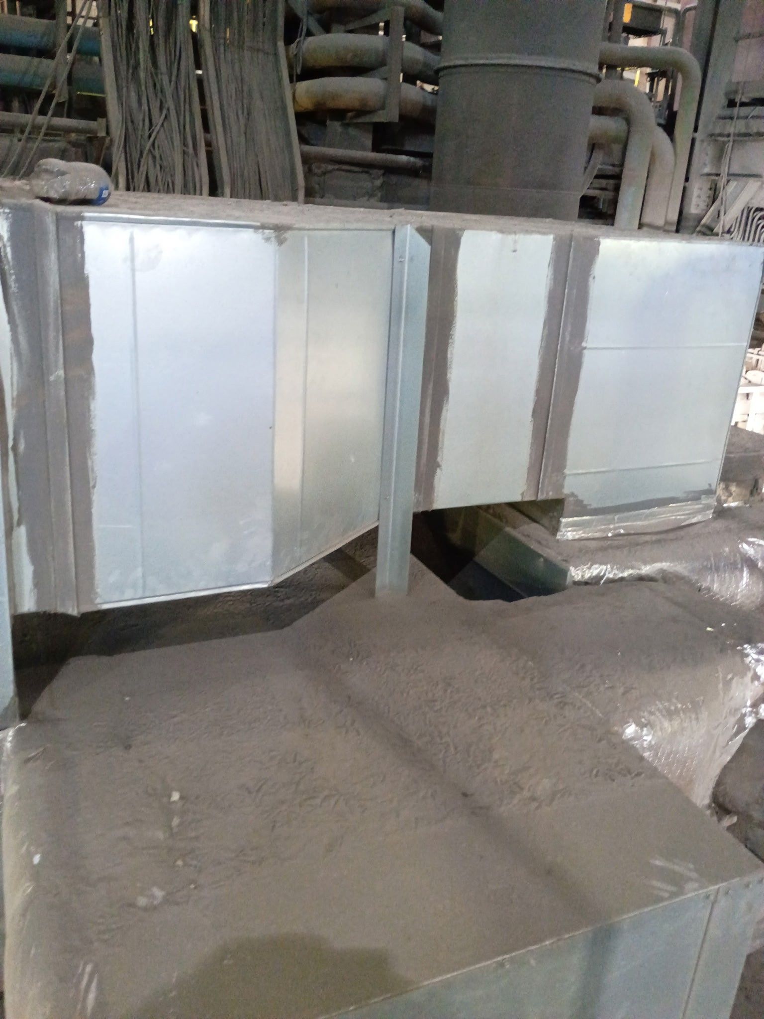 Galvanized metal ductwork in an industrial setting, with sealed seams and surrounding floor covered in fine, dark material.