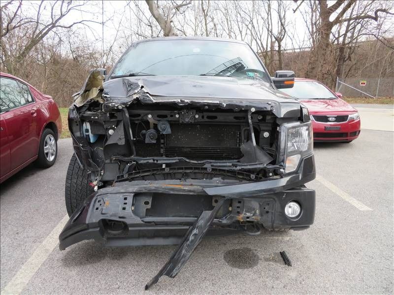 Damaged black Ford truck after a collision, with a crushed front end. Two other cars are visible.