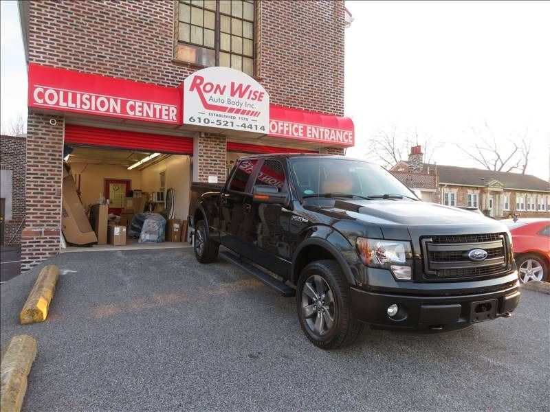 Black Ford pickup truck parked in front of Ron Wise Auto Body Collision Center; red brick building.