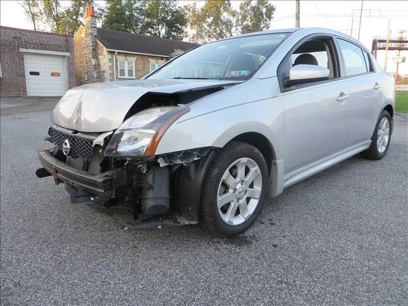 Silver Nissan Sentra with front-end damage, parked in a lot. Building in the background.