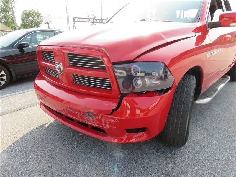Red Dodge Ram pickup truck with front-end damage. Cracked bumper, headlight and hood damage; parked on asphalt.