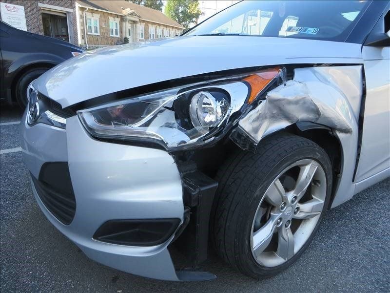 Silver car with front-end damage; crumpled fender, headlight intact, tire exposed, parked outdoors.