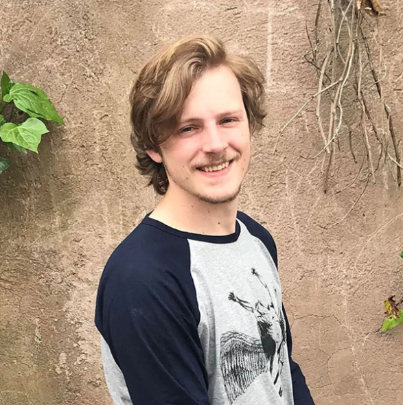 Man with light brown hair smiles, wearing a navy and gray raglan shirt, standing against a textured wall with foliage.