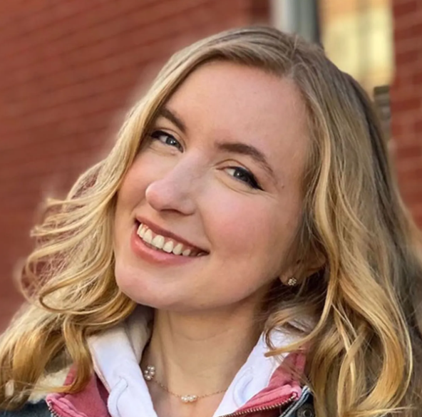 Blonde woman smiling, wearing a pink jacket, against a red brick building.