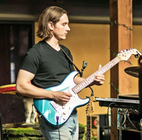 Guitarist playing a blue electric guitar outdoors. He's wearing a black shirt and jeans, focused on his instrument.