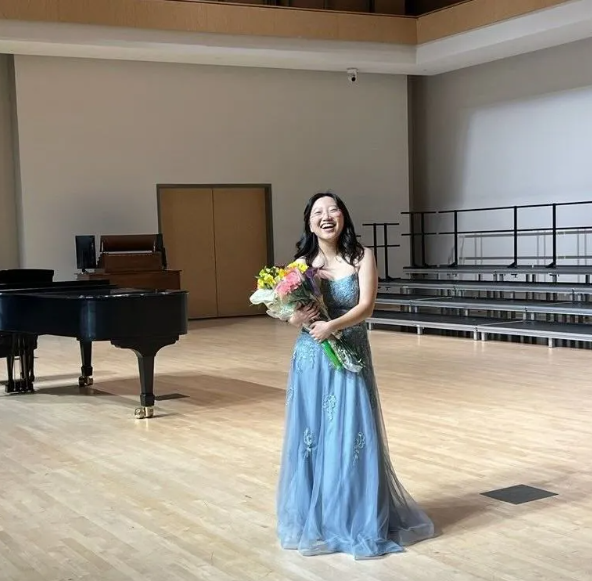 Woman in blue dress holds flowers, smiles, stands in empty hall with piano.