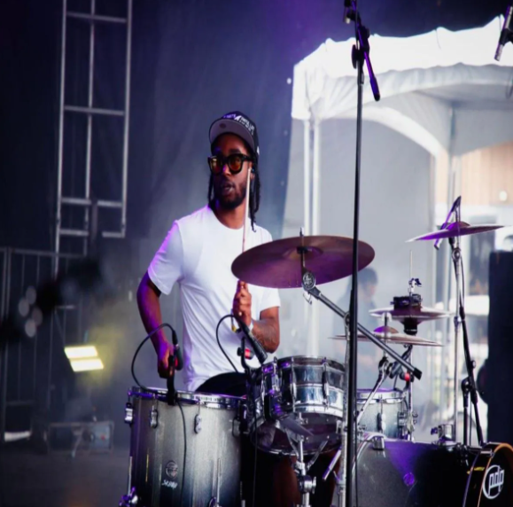 Drummer playing on stage, wearing headphones and a white shirt. Stage lit with purple lights.