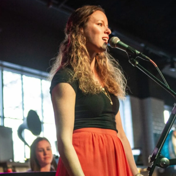 Woman singing into a microphone on stage, wearing a black top and orange skirt.