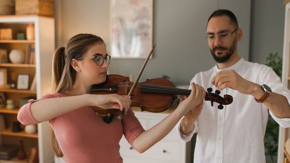 A person with glasses plays a violin while a teacher adjusts the tuning pegs in a brightly lit room.