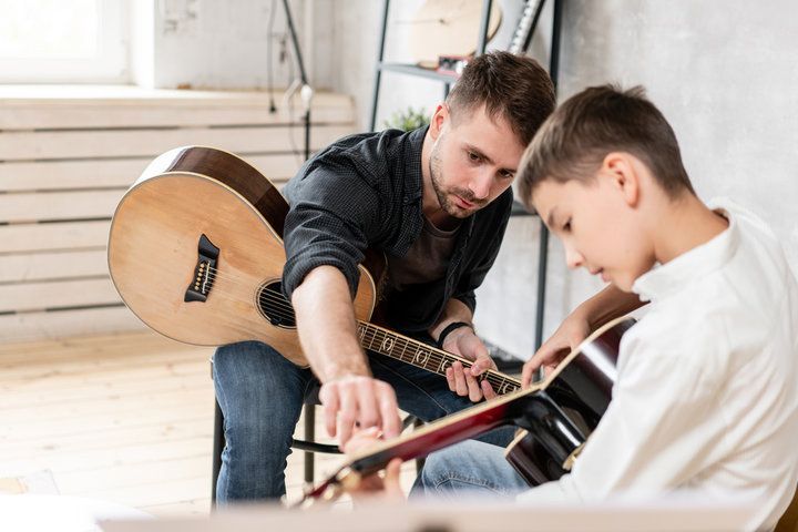 An instructor guides a student's hand on the fretboard of an acoustic guitar during a music lesson.