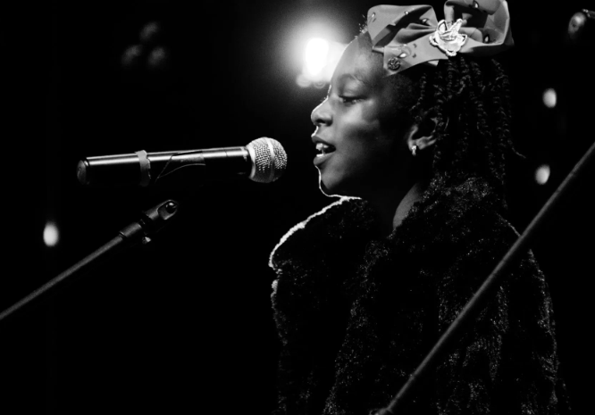 Young person singing into a microphone on stage, wearing a bow and fur jacket.