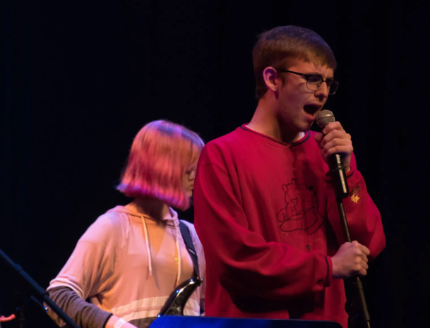 Man in red sweater singing into microphone on stage; guitarist in background.