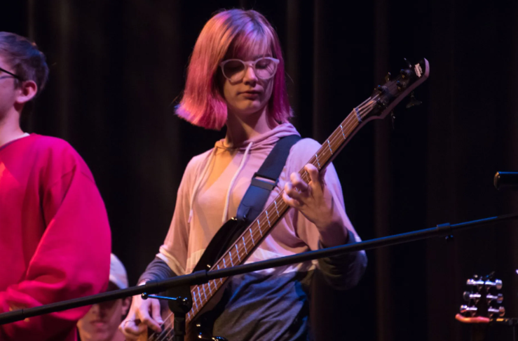 Person with pink hair plays bass guitar on stage, wearing glasses and a light-colored hoodie.