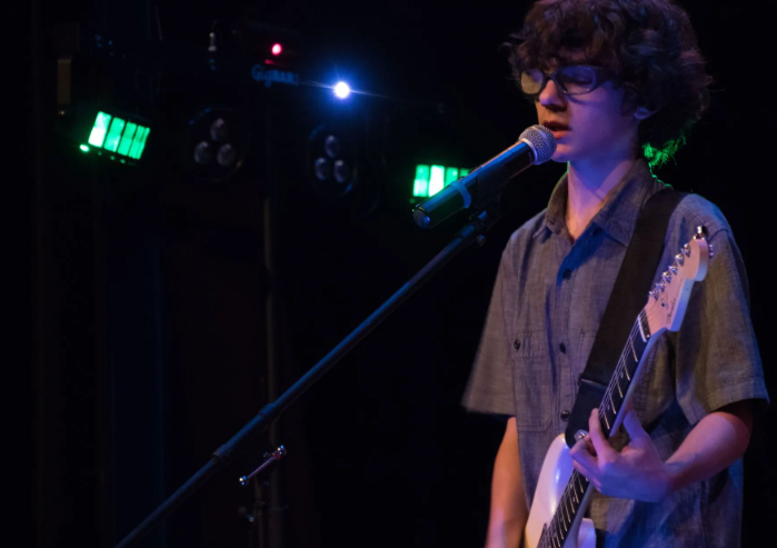 Person on stage singing and playing a white electric guitar, under green and blue stage lights.