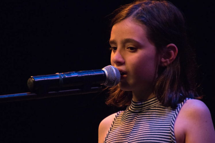 Girl singing into a microphone on stage, wearing a striped top, with dark background.