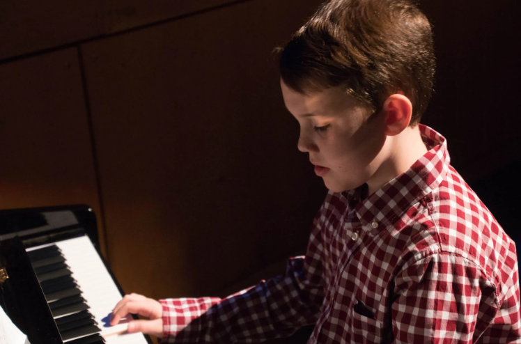Boy in red plaid shirt playing piano, lit from the side.