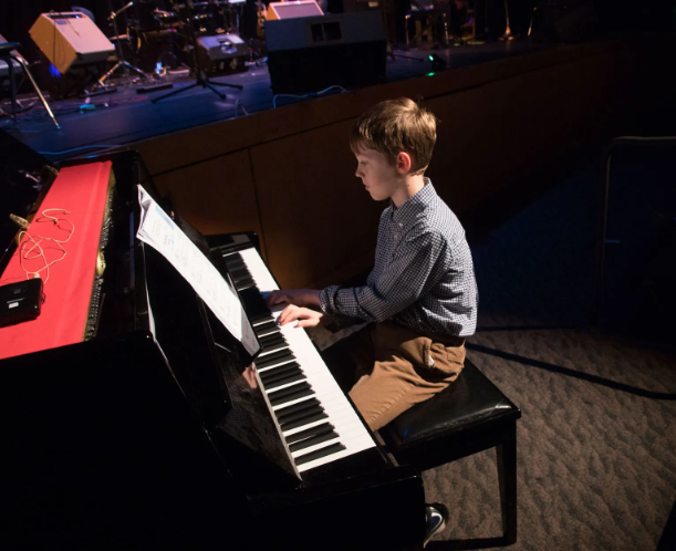 Young person playing a piano on a stage, focused expression, sheet music present.